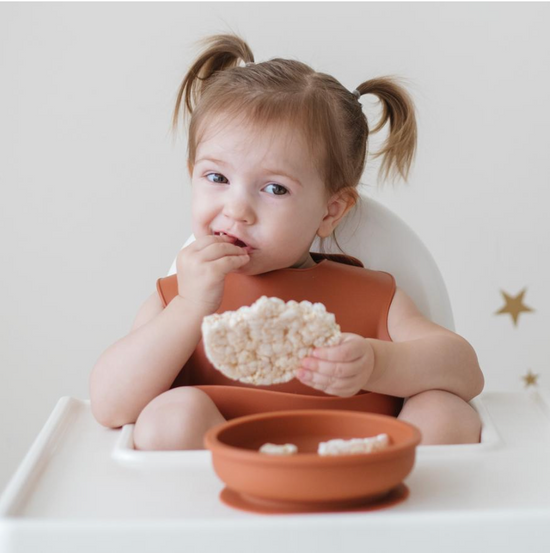 young girl eat rice cake in high chair with rust-coloured bib and plate